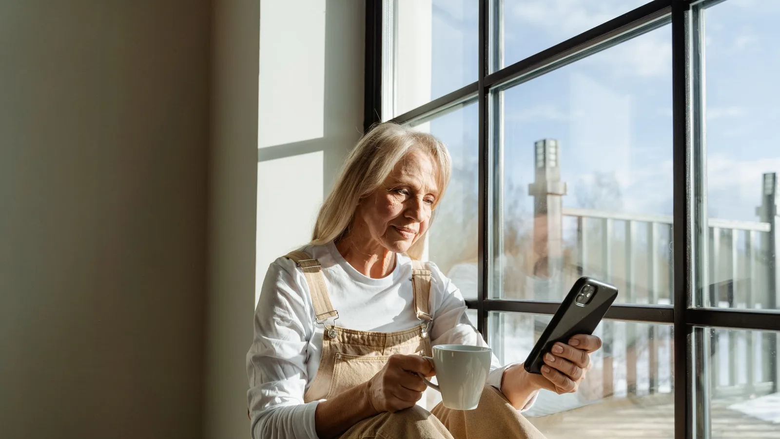 A woman in her sixties with a coffee and her phone by a sunny window